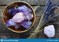A bowl of purple quarts. Different shades of purple. Lavender Stalks tied with twine. Laid down next to the bowl of purple quartz. Everything sits on a wooden table.